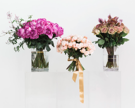 Pink and purple roses in three glass vases on a clear podium.